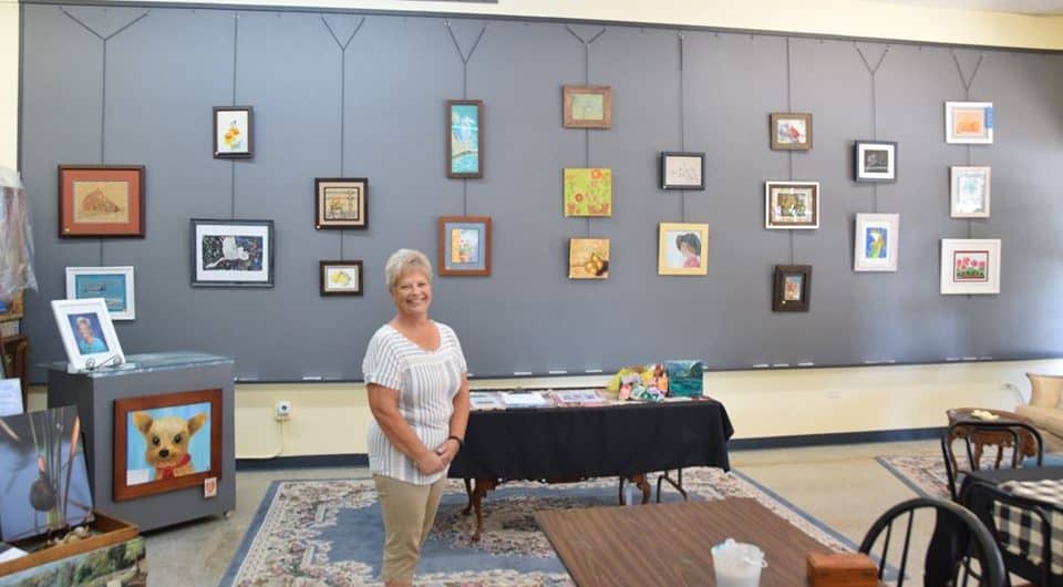 Vicki Alley smiling in the Gray Gallery in front of a wall of her artworks.
